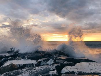 Sea waves splashing on rocks against sky during sunset