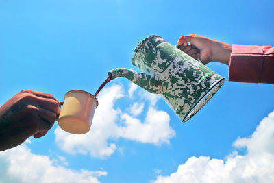 Low angle view of woman pouring coffee in person holding cup against blue sky