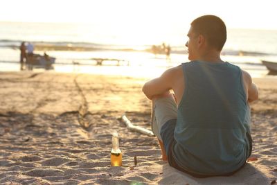 Rear view of young man with beer bottle sitting on beach against sky during sunset
