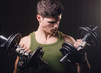 Young man exercising with dumbbells while standing against black background