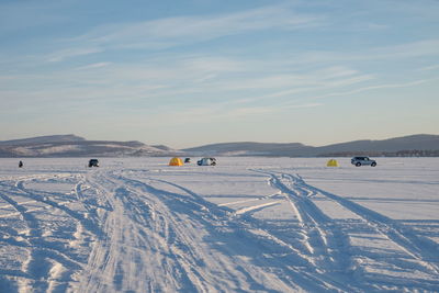 Car tracks lead to fishermen who catch fish on the lake, against mountains, in winter.