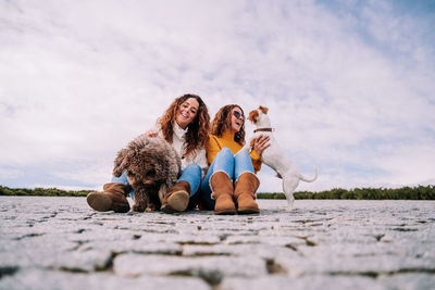 Smiling women playing with dog sitting against sky