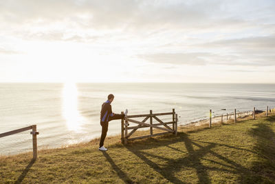 Full length of man stretching leg on fence at beach during sunny day