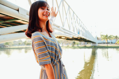 Portrait of smiling young woman standing on bridge over river