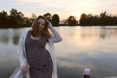 Portrait of pregnant woman standing by lake against sky during sunset