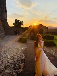 Woman standing in evening gown on road against sky during sunset