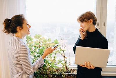 Smiling female colleagues standing with technologies by window at creative office