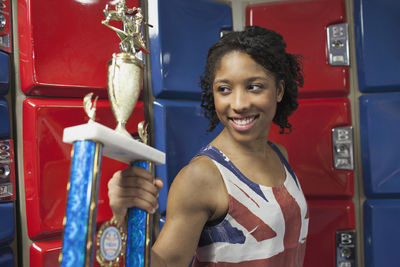 A young woman with a trophy in fornt of red and blue lockers.