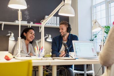 Cheerful multi-ethnic business colleagues sitting at desk with laptop and microphone in creative office