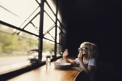 Girl wearing eyeglasses having food while sitting at table by window in darkroom