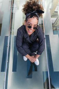From above of laughing african american female in trendy outfit sitting on glass roof and embracing knees while looking at camera