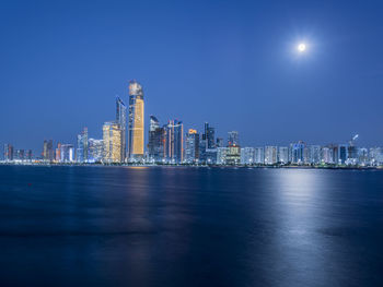 Illuminated buildings by sea against sky at night