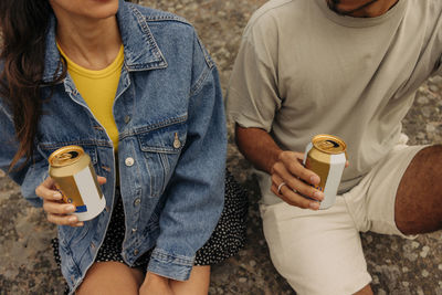 Midsection of male and female friends holding drink cans while sitting together on rock