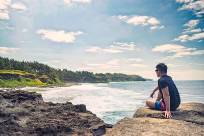 Man sitting on rocky shore against blue sky