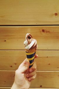 Cropped hand of woman holding ice cream cone against wooden wall