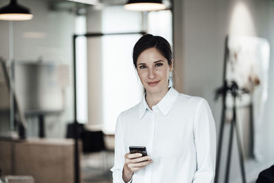 Confident businesswoman holding smart phone while standing in office