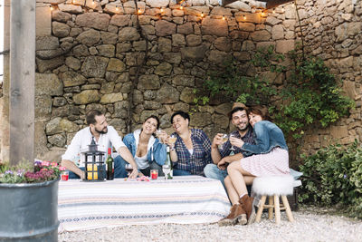 Happy friends with wine partying in yard against stone wall during sunset