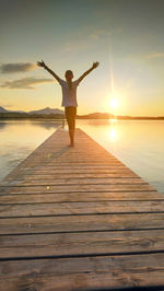 Rear view of woman with arms outstretched standing on pier over lake during sunset