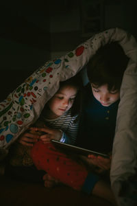 Siblings using digital tablet on bed in darkroom