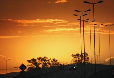 Silhouette trees by street against orange sky during sunset