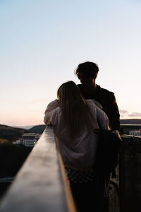 Couple standing on footbridge against sky during sunset