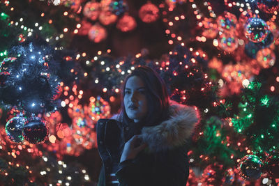 Thoughtful young woman standing against illuminated christmas tree at night