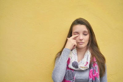 Young woman rubbing eye while standing against yellow wall