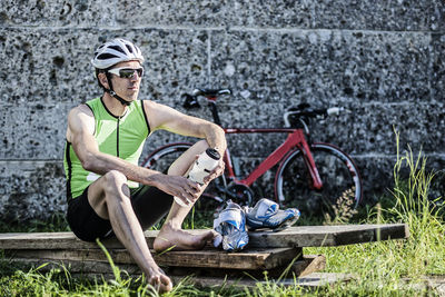 Cyclist holding water bottle while relaxing on wooden plank against wall