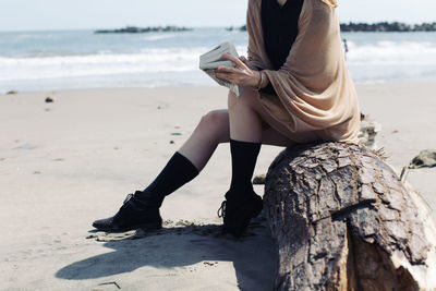 Low section of woman sitting with book on log at beach