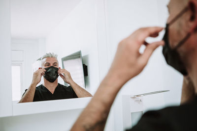 Serious male tattoo master standing in salon in front of mirror and putting on protective mask before procedure