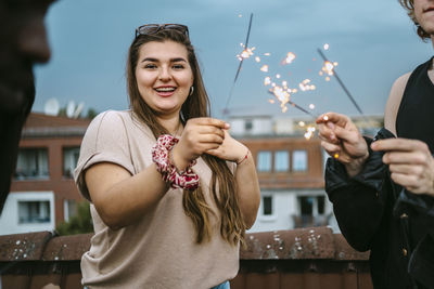 Portrait of smiling young woman holding sparklers with male friends on rooftop