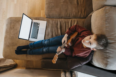 High angle portrait of boy playing guitar while sitting on sofa at home