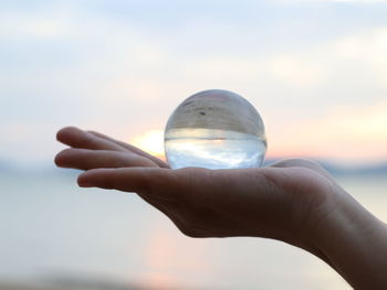 Close-up of hand holding crystal ball against sky during sunset