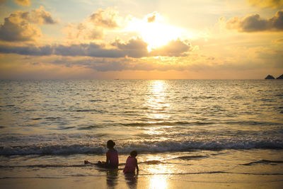 Girls playing on shore at beach against cloudy sky during sunset