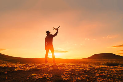 Silhouette man with toy airplane standing on land against sky during sunset