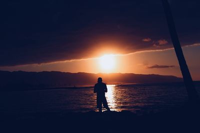 Silhouette man standing in sea against sky during sunset