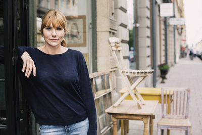 Portrait of woman standing by store against chairs on sidewalk in city