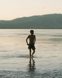 Rear view of man standing in sea against sky during sunset