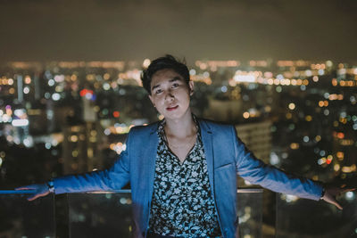 Portrait of young man standing against illuminated cityscape at night