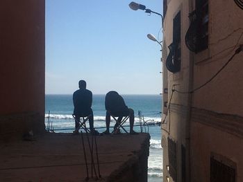 Rear view of men sitting on chair by sea against sky