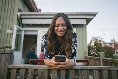 Smiling mid adult woman using mobile phone while leaning on railing at porch