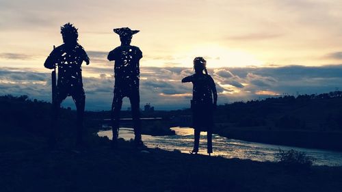 Silhouette people on riverbank against cloudy sky during sunset