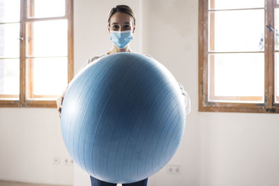 Woman wearing face mask holding fitness ball while standing at office