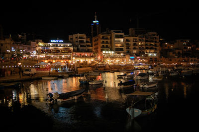 Boats moored at harbor against illuminated buildings in city at night