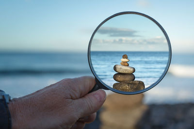 Cropped hand holding magnifying glass in front of pebbles stack at beach