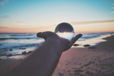 Close-up of hand holding crystal ball at beach against sky during sunset