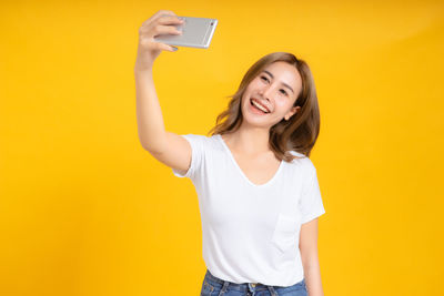 Smiling young woman photographing while standing against yellow background