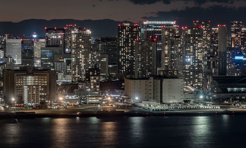 Illuminated modern buildings by river against sky at night