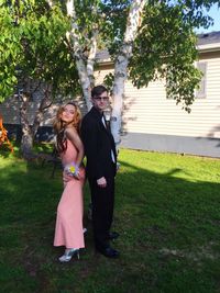 Full length of cheerful teenage couple standing on grassy field at yard during prom
