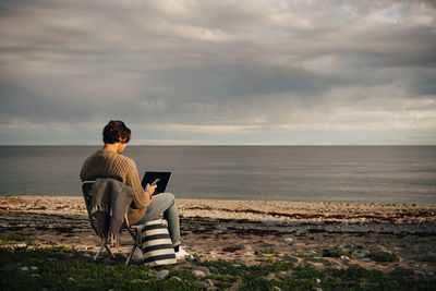 Mid adult man using laptop and mobile phone while sitting on sea shore at beach against sky